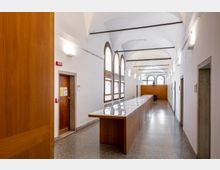 Corridoio interno di un edificio istituzionale con soffitto a volte, pareti bianche e pavimento in graniglia. Lungo un lato ci sono grandi finestre ad arco e al centro una lunga fila di banconi vetrati; in fondo si vede una parete in legno con la scritta “RETTORATO”.