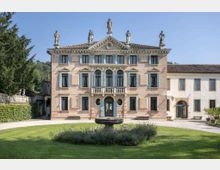 Facciata di una villa storica a tre piani con frontone centrale, statue sul tetto, finestre simmetriche e balcone con balaustra sopra l’ingresso ad arco. Davanti si vede un prato curato con una fontana circolare, mentre a destra è collegato un edificio annesso più semplice.