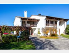 Casa indipendente a due piani con facciata bianca, tetto a falde e balconi in legno, affacciata su un cortile con vialetto in ghiaia. Davanti si vedono giardino con prato, una pergola coperta di rampicanti e alberi ornamentali sotto un cielo sereno.