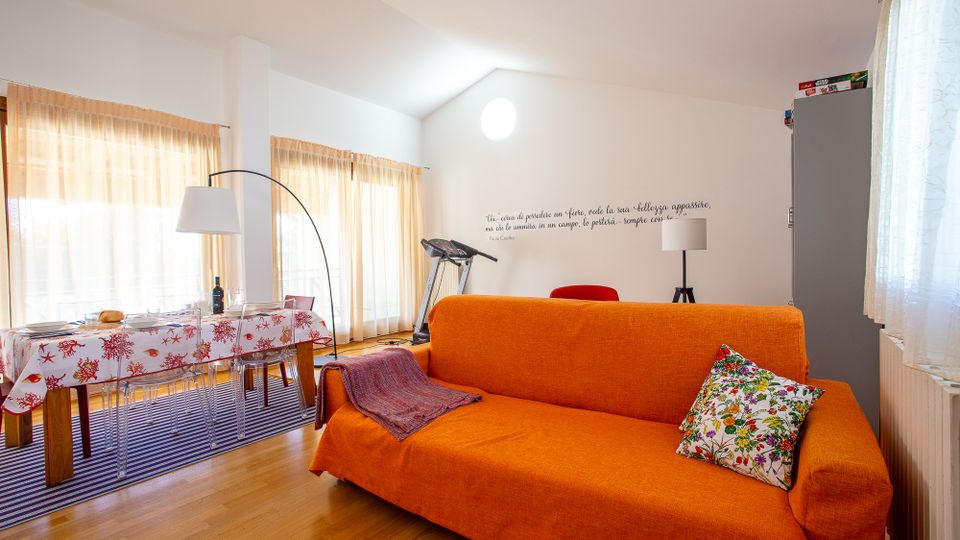 Soggiorno open space luminoso con pavimento in legno, divano arancione in primo piano e zona pranzo con tavolo e sedie trasparenti vicino a grandi finestre con tende chiare. Sullo sfondo si vedono una lampada ad arco, una cyclette e pareti bianche con soffitto inclinato e finestra circolare.