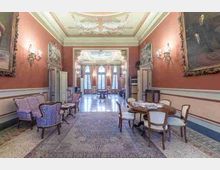 Salone storico con pareti rosse, grandi quadri incorniciati e applique dorate, pavimento in marmo con grande tappeto centrale e arredi d’epoca (poltrone e tavolo). In fondo un’ampia apertura conduce a una sala luminosa con alte finestre e tende, sotto un soffitto decorato con stucchi e affreschi.