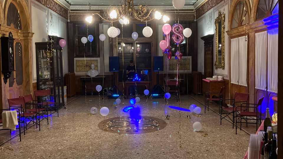 Salone interno in stile storico con soffitto a travi dipinte, lampadario centrale e pareti decorate con archi e specchi dorati. La sala ha pavimento in terrazzo con medaglione centrale ed è allestita per una festa con palloncini, luci colorate e postazione audio sul fondo.