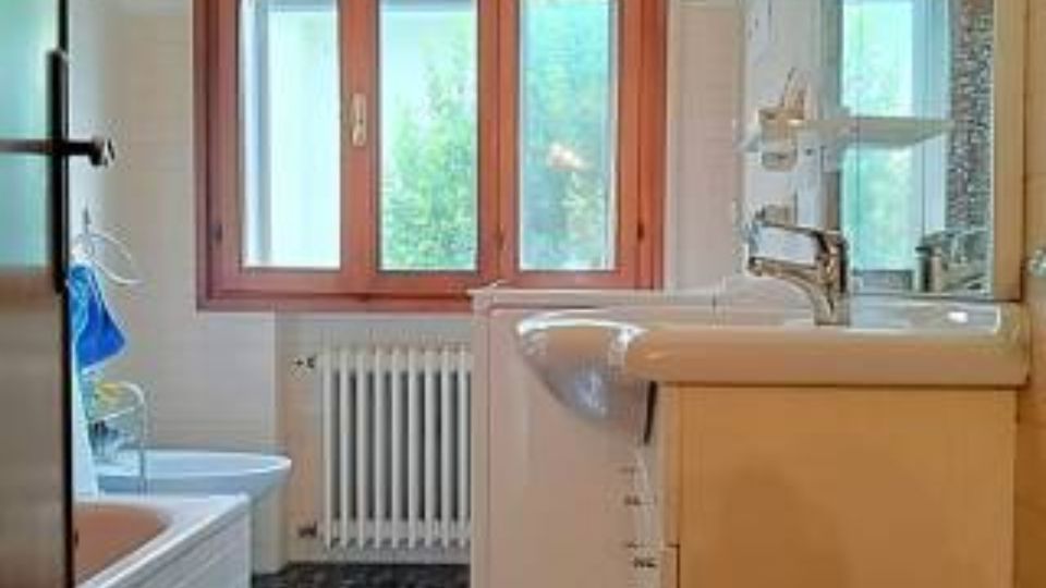 Bagno domestico luminoso con grande finestra a tre ante con telaio in legno e radiatore sotto. In primo piano si vedono il lavabo con mobile e specchio, mentre a sinistra è presente una vasca e il pavimento è rivestito con piastrelle scure.