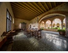 Loggiato di un chiostro storico con soffitto a travi in legno, pavimento in pietra e un grande tavolo con sedie in legno. Sul lato aperto si vedono archi in pietra che affacciano su un cortile interno con aiuole e vasi di piante.