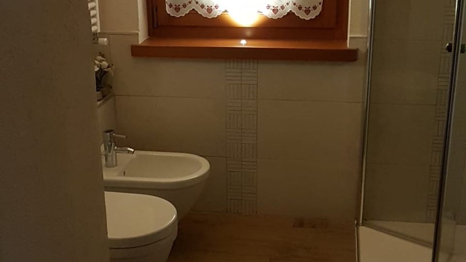 Bagno moderno con pavimento effetto legno, sanitari bianchi sospesi e box doccia in vetro sulla destra. Sul fondo c’è una finestra con cornice in legno e tendina bianca, sopra pareti rivestite con piastrelle chiare.