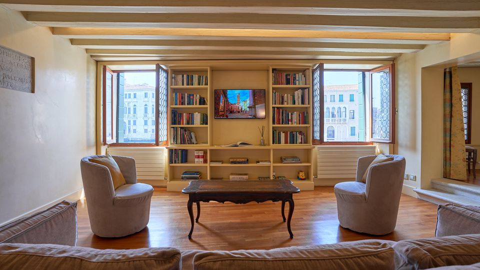 Soggiorno luminoso con pavimento in legno e soffitto a travi a vista, arredato con poltrone e un tavolino centrale di fronte a una libreria a parete con TV. Due grandi finestre aperte incorniciano la vista su edifici storici all’esterno.
