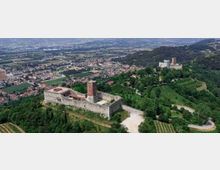 Veduta aerea di una fortezza medievale in pietra con mura rettangolari e una torre centrale, circondata da boschi e colline. Sullo sfondo si estendono vigneti e un’area urbana con campi nella pianura.