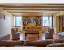 Soggiorno luminoso con pavimento in legno e soffitto a travi a vista, arredato con poltrone e un tavolino centrale di fronte a una libreria a parete con TV. Due grandi finestre aperte incorniciano la vista su edifici storici all’esterno.