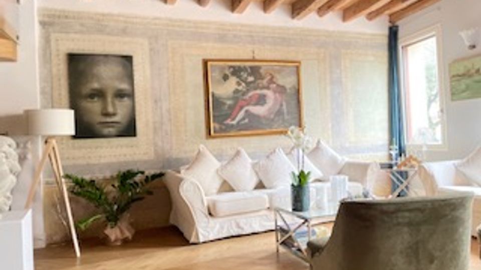 Soggiorno luminoso con soffitto a travi in legno, pareti chiare e pavimento in legno. Divano bianco con cuscini, tavolino in vetro, lampada da terra, quadri alle pareti e grandi finestre che illuminano l’ambiente.