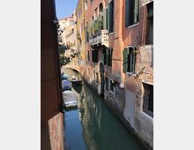 Canale stretto tra alti edifici storici con facciate in intonaco e mattoni, persiane verdi e balconi in pietra affacciati sull’acqua. Piccole barche sono ormeggiate lungo la riva e un ponte ad arco in muratura attraversa il canale sullo sfondo.