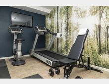 Piccola sala palestra con cyclette e tapis roulant davanti a una parete blu con TV a schermo piatto. Sulla parete laterale c’è una grande carta da parati con bosco illuminato dal sole, e in primo piano una panca regolabile con manubri su pavimento piastrellato.