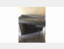 Interno di un edificio con una stretta scala in legno che scende in un’apertura rettangolare nel pavimento. Pareti intonacate chiare e gradini consumati definiscono un accesso a un livello inferiore buio.