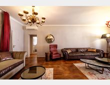 Soggiorno interno con pavimento in legno, divani in pelle e poltrona rossa, tavolini bassi e lampadario in ottone. Pareti chiare con specchio rotondo, tenda rossa a tutta altezza e porta d’ingresso con apertura verso un corridoio sullo sfondo.