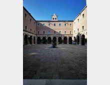 Cortile interno di un edificio storico in mattoni, con ampio pavimento in pietra e porticato ad arcate su tre lati. Al centro si vede una piccola fontana e sul lato opposto una facciata con finestre regolari e un piccolo frontone con orologio sotto un cielo limpido.