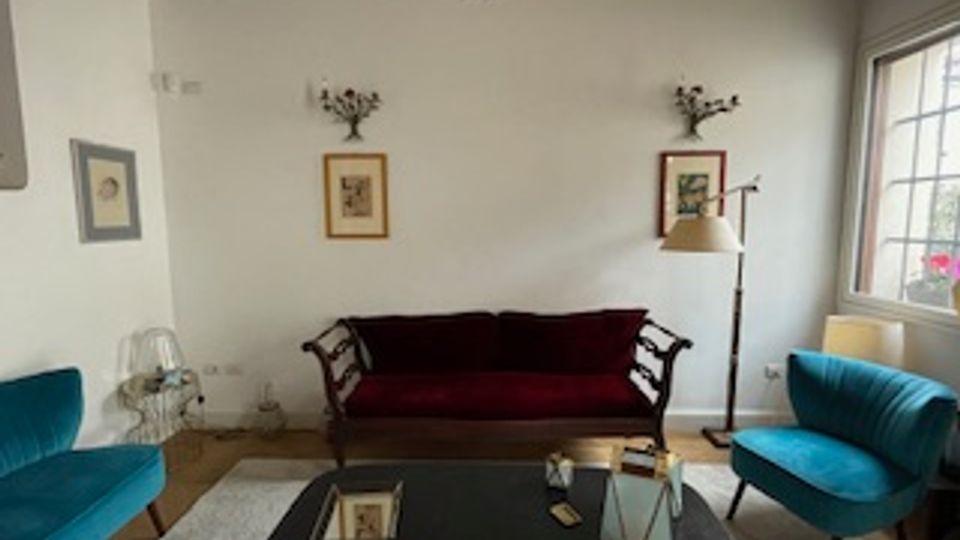 Salotto con soffitto a travi in legno a vista, pareti bianche con quadri e divano scuro al centro, affiancato da due poltrone turchesi. Davanti c’è un tavolino da caffè con oggetti decorativi e, sulla destra, una grande finestra con persiane che illumina l’ambiente.