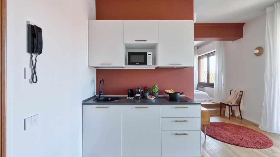 Angolo cucina moderno in un monolocale, con mobili bianchi, piano di lavoro scuro, lavello nero e microonde incassato su parete rosso mattone. A destra si intravede la zona giorno/notte con finestra, tende bianche, pavimento in legno e tappeto rotondo rosso.