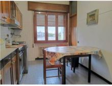 Cucina stretta con mobili in legno e piano cottura sul lato sinistro, pavimento piastrellato e tavolo rettangolare con tovaglia al centro. In fondo una grande finestra con persiane e radiatore sotto, con pareti chiare e un quadro appeso.