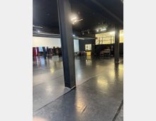 Ampio spazio interno tipo sala prove o studio teatrale con pavimento nero lucido e grandi colonne metalliche. Dal soffitto scuro pendono fari e tubazioni, con un soppalco laterale e alcune attrezzature e sedie lungo le pareti.