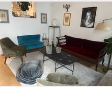 Soggiorno con pareti bianche arredato con divano bordeaux, divanetto blu e poltrone, centrato su un tavolino basso in metallo su tappeto chiaro. Alle pareti sono appesi quadri e una lampada da terra illumina l’angolo vicino a una pianta.