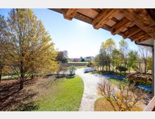 Vista da un portico con travi in legno su un giardino con prato, vialetto curvo e cancello bianco, circondato da alberi. Sullo sfondo si vedono campi coltivati e alcune case in una zona rurale.