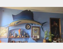 Parete interna dipinta di azzurro con un grande pesce vela tassidermizzato montato in alto e una nicchia con mensole piene di oggetti decorativi, cornici e strumenti. Ai lati si vedono quadri incorniciati, un medaglione al centro e alcune piante in vaso su un mobile.