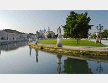 Veduta di un lungofiume con un’ampia area verde affiancata da un canale calmo che riflette il cielo e le statue in pietra. Sullo sfondo si vedono edifici storici e una grande basilica con cupole e campanili.