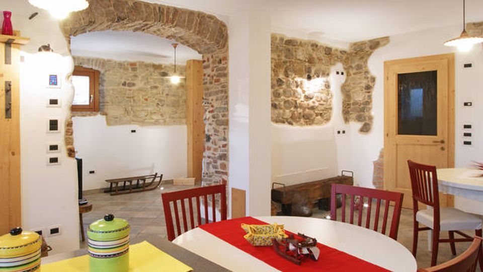 Interno di una cucina-sala da pranzo con pareti bianche e inserti in pietra a vista, arco in muratura e pavimento in piastrelle. Tavolo con runner rosso e sedie in legno, piano cucina con accessori, porta in legno e lampade a sospensione.