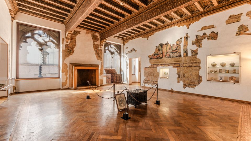 Sala museale in un palazzo storico con pavimento in parquet a spina di pesce e soffitto a travi lignee decorate. Alle pareti, intonaco parzialmente scrostato con affreschi visibili, grandi finestre ad arco, un camino in pietra e vetrine illuminate con ceramiche esposte.