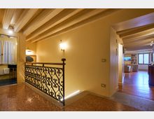 Interno di una casa con soffitto a travi in legno e pareti color crema, con corrimano in ferro battuto lungo un piccolo ballatoio su pavimento in terrazzo. A destra si apre un soggiorno con pavimento in legno, libreria e grande finestra, mentre a sinistra si intravede un bagno con finestra e tende bianche.