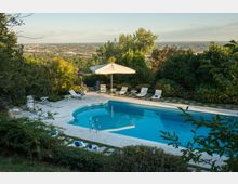 Piscina all’aperto con bordo in pietra chiara e acqua azzurra, circondata da siepi e alberi su un pendio. Intorno ci sono lettini e un grande ombrellone, con vista panoramica sulla pianura e su un’area urbana in lontananza.