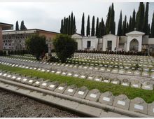 Cimitero con file ordinate di lapidi basse in pietra su aiuole erbose e vialetti di ghiaia, con fiori e piccoli monumenti. Sullo sfondo si vedono cappelle e loculi, con alti cipressi che delimitano l’area.