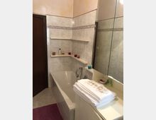 Bagno con vasca incassata lungo la parete, rivestimenti in piastrelle chiare con fascia decorativa e mensole angolari. A destra c’è un grande specchio sopra un piano d’appoggio con asciugamani piegati e piccoli contenitori, e a sinistra una porta in legno scuro.