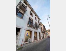 Facciata di un edificio residenziale a più piani lungo una strada stretta, con intonaco chiaro, finestre con scuri e piccoli balconi in ferro battuto. Al piano terra si vedono vetrine e ingressi, un’unità esterna di climatizzazione e dissuasori rossi sul marciapiede, con lampione e altri edifici allineati lungo la via.