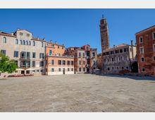 Ampia piazza lastricata in pietra circondata da edifici storici a più piani con facciate color pastello e finestre ad arco. Sullo sfondo si innalza un alto campanile in mattoni sopra i tetti in coppi, sotto un cielo sereno.