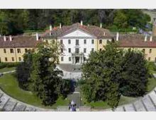 Veduta frontale di una grande villa storica simmetrica con facciata chiara e tetto in tegole, affiancata da due ali laterali. Davanti si trova un cortile con viali e aiuole, circondato da alberi maturi e aree verdi.