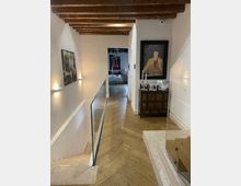 Corridoio interno di una casa con pareti bianche, soffitto con travi in legno a vista e pavimento in parquet a spina di pesce. Lungo le pareti ci sono applique, quadri e una credenza con un grande dipinto, con un’apertura verso un’altra stanza in fondo.