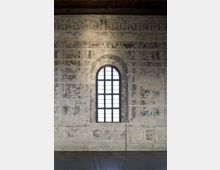 Interno di un edificio storico con grande finestra ad arco a griglia al centro e pareti intonacate decorate da affreschi molto consumati. In alto si vede il soffitto con travi in legno e in basso un pavimento liscio in pietra.