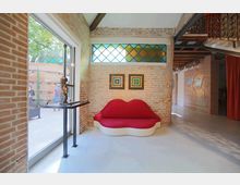 Interno di uno spazio espositivo in stile industriale con pareti in mattoni a vista, pavimento chiaro e travi in legno, illuminato da una grande porta vetrata. Al centro c’è un divano rosso a forma di labbra sotto una finestra con vetri colorati, con quadri alle pareti e un soppalco con ringhiera in ferro battuto.