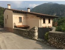 Casa rurale con facciata beige decorata, tetto con tegole rosse, e un piccolo portico, situata lungo una strada asfaltata. Sullo sfondo si intravedono colline boscose e una recinzione in pietra delimita il cortile.