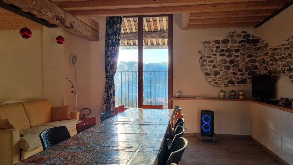 Interno di una sala da pranzo rustica con soffitto in travi di legno, lungo tavolo con sedie e un divano laterale. Una grande porta finestra conduce a un balcone con vista sulle montagne, mentre una parete in pietra a vista e un mobile con TV completano l’ambiente.