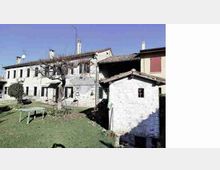 Cortile erboso in un contesto rurale con edifici in muratura intonacata e pietra, a due piani, con tetti in coppi e camini. In primo piano si vede un piccolo annesso in pietra con tetto spiovente e un tavolo all’aperto vicino a un albero spoglio.