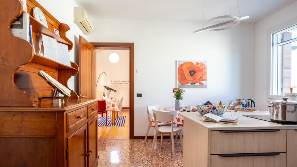 Cucina-sala da pranzo luminosa con pavimento in terrazzo, grande credenza in legno a sinistra e isola moderna con cassetti in primo piano. Tavolo con sedie sotto una finestra, lampada a sospensione circolare e quadro floreale sulla parete, con porta aperta verso un’altra stanza.