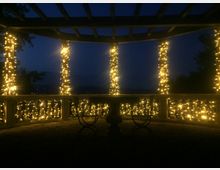 Gazebo o pergolato all’aperto visto di notte, con travi in legno e colonne avvolte da luci a stringa. Una ringhiera decorativa illuminata circonda lo spazio, con sagome di sedute in primo piano e cielo scuro sullo sfondo.