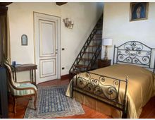 Camera da letto in stile classico con letto in ferro battuto, pavimento in legno e tappeto decorato. Sullo sfondo si vedono una porta bianca, un comodino con lampada e una scala interna in metallo e legno che conduce a un soppalco.