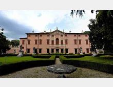 Facciata di una villa storica color rosa con portico centrale e frontone, affacciata su un giardino formale con siepi geometriche e statue. In primo piano si vede un vialetto centrale e una piattaforma circolare in pietra su ghiaia.