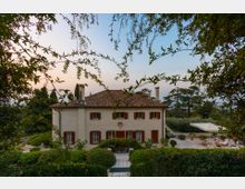 Villa di campagna a due piani con facciata chiara, tetto in coppi e persiane rosse, circondata da giardini curati e siepi. In primo piano si vede un’area esterna pavimentata con ombrellone e sedute, immersa nel verde di alberi e arbusti.