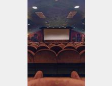 Interno di una sala cinematografica con file di poltrone imbottite rivolte verso un grande schermo bianco. Soffitto curvo con luci circolari incassate, pareti laterali con rivestimenti scuri e apparecchiature ai lati dello schermo.