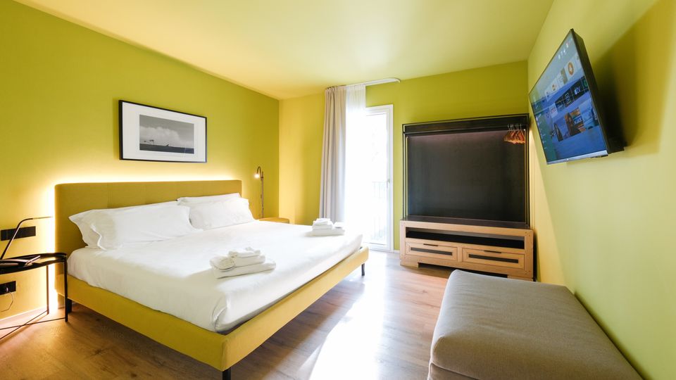 Camera da letto moderna con pareti verde chiaro e pavimento in legno, con letto matrimoniale e comodino. Di fronte ci sono una TV a parete, un mobile contenitore con appendiabiti e una porta-finestra con tende che illumina l’ambiente.