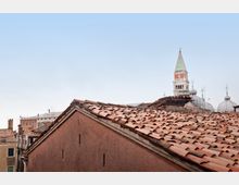 Veduta sui tetti in tegole di terracotta di edifici storici, con un campanile slanciato e alcune cupole sullo sfondo. Cielo sereno sopra il centro urbano.
