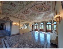 Ampio salone in stile storico con soffitto a cassettoni riccamente decorato in stucco e affreschi, pareti con grandi specchi e applique, e pavimento in terrazzo a motivi geometrici. Sul fondo, alte portefinestre ad arco lasciano entrare luce naturale e si affacciano su un panorama esterno.